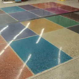 Wielobarwna, błyszcząca posadzka z segmentów w kształcie kwadratów, wykonana z materiału z widocznymi drobnymi ziarnami, z odbiciami światła. Widoczny sprzęt do polerowania w tle.