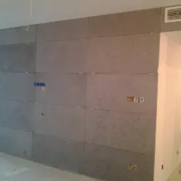 Ściana wykończona płytami betonowymi w szarym odcieniu z widocznymi gniazdkami elektrycznymi i puszkami instalacyjnymi, drabina aluminiowa oparta o ścianę w tle.