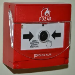Czerwony, naścienny przycisk alarmowy firmy Polon-Alfa z instrukcją aktywacji: 'Uderz w szybkę, wciśnij przycisk'. Symbol domu w płomieniach.