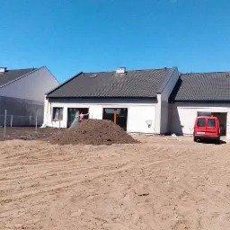 Osiedle nowych, parterowych domów jednorodzinnych z szarymi dachami i białymi elewacjami, widok zza szyby pojazdu na plac budowy z pryzmą ziemi i czerwonym samochodem dostawczym.