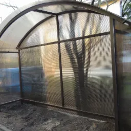 Zadaszona wiata z poliwęglanu o metalowej konstrukcji, widok z zewnątrz, przez przezroczyste ściany widać zarys krzewów i drzew.