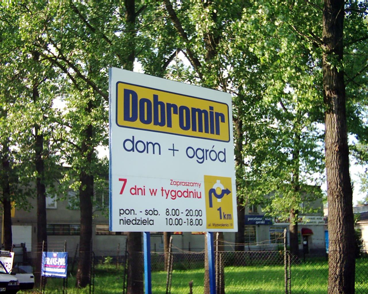 Tablica reklamowa firmy Dobromir oferującej usługi dla domu i ogrodu, z informacją o godzinach otwarcia i odległości 1km, umieszczona na tle drzew i ogrodzenia.