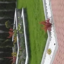 Kaskadowy ogród z trawnikiem, rabatami kwiatowymi w białych obrzeżach i ścieżką z czerwonej kostki brukowej, widok z góry.