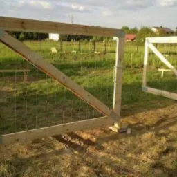 Surowa, drewniana brama ogrodowa z metalową siatką, w trakcie montażu, z widocznymi słupkami i konstrukcją wzmacniającą, na tle zielonej trawy i zabudowań.