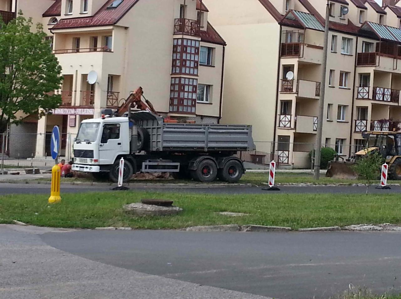 Ciężarówka wywrotka z dźwigiem HDS na tle budynków mieszkalnych, widoczny fragment wykopu zabezpieczonego barierkami, żółto-czarny pachołek i znaki drogowe.
