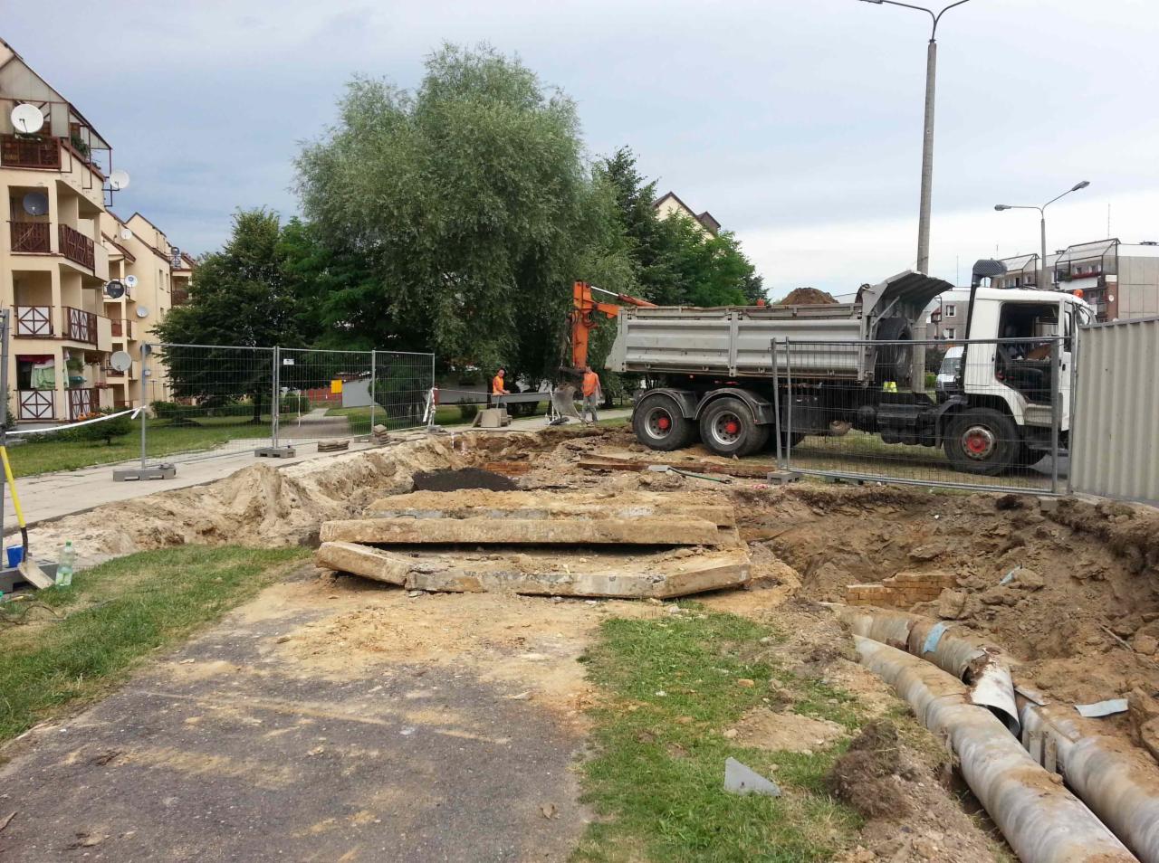 Wykop w terenie miejskim z widocznymi rurami, zabezpieczony ogrodzeniem tymczasowym, z koparką załadowującą ziemię na wywrotkę. W tle budynki mieszkalne i drzewa.