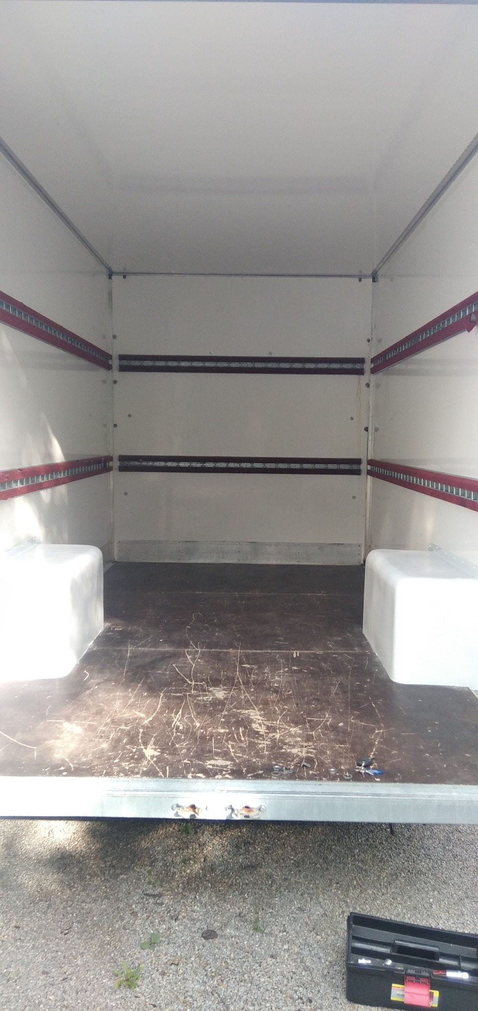 Pusta przestrzeń ładunkowa busa z drewnianą podłogą zarysowaną śladami użytkowania, widoczne mocowania na ścianach i skrzynka z narzędziami na zewnątrz pojazdu.