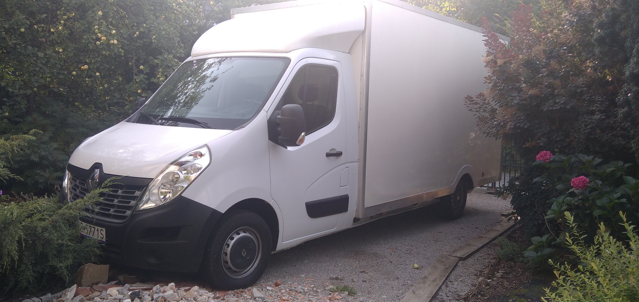 Biały samochód dostawczy Renault Master z zabudową kontenerową zaparkowany na żwirowej drodze, otoczony zielenią krzewów i drzew, z widocznymi różowymi kwiatami hortensji.