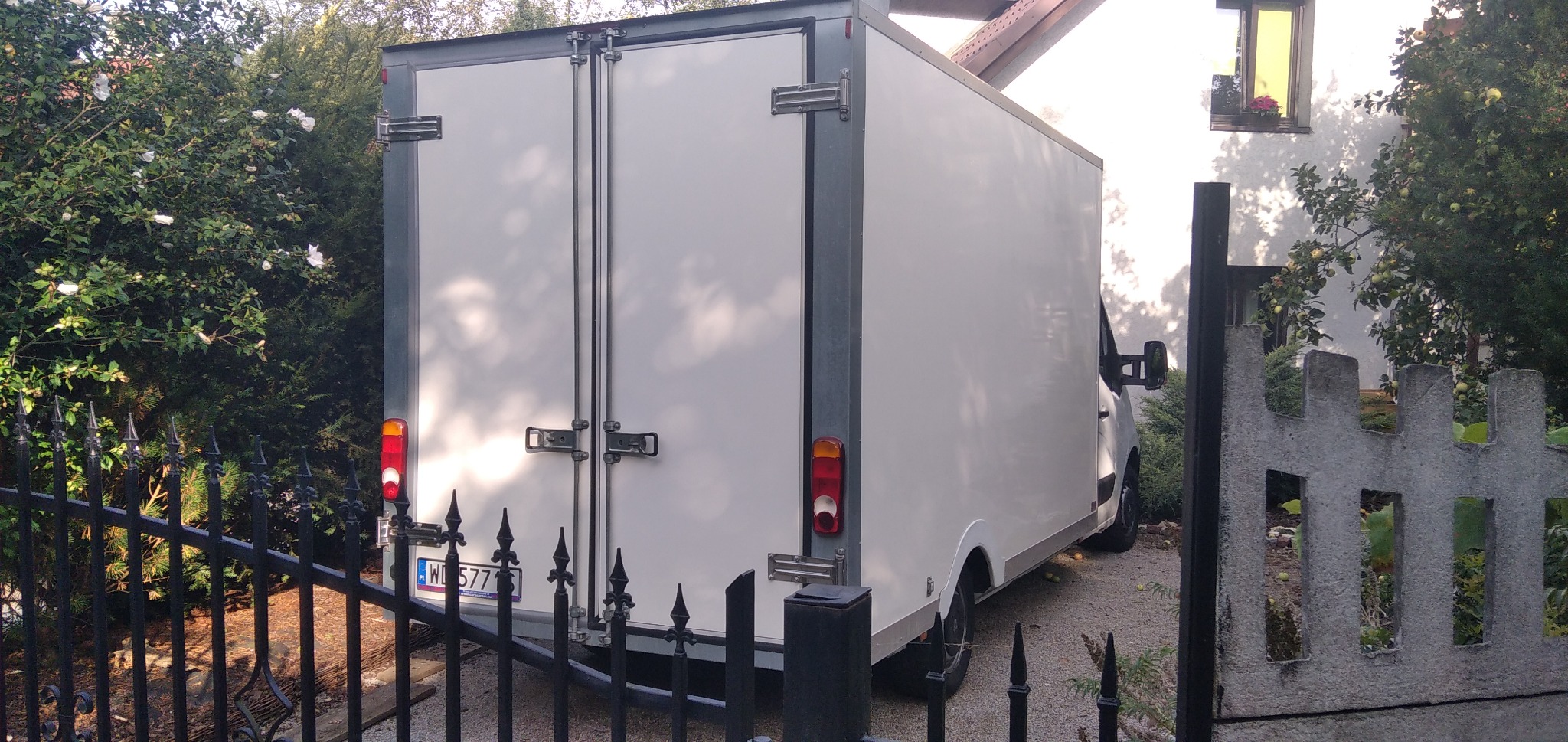 Tył białego furgonetki z zamkniętymi drzwiami, zaparkowanego na podwórku z widoczną tablicą rejestracyjną przez czarny, ozdobny płot.