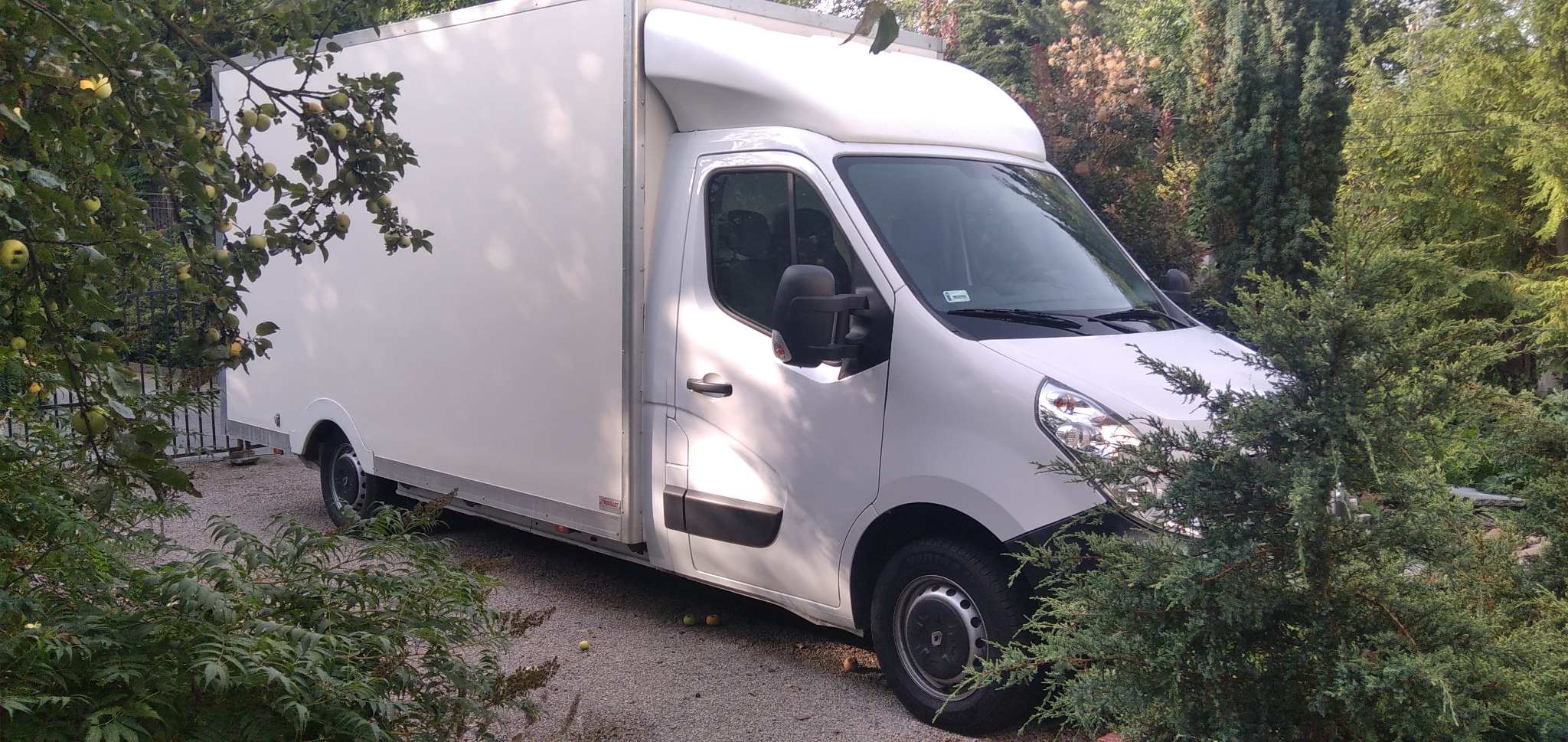 Biały samochód dostawczy Renault Master zaparkowany na żwirowej drodze w otoczeniu zieleni drzew i krzewów. Na drzewie po lewej stronie widoczne zielone jabłka.
