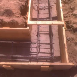 Zbrojenie fundamentów w drewnianym szalunku, widoczne pręty stalowe i piasek w tle.