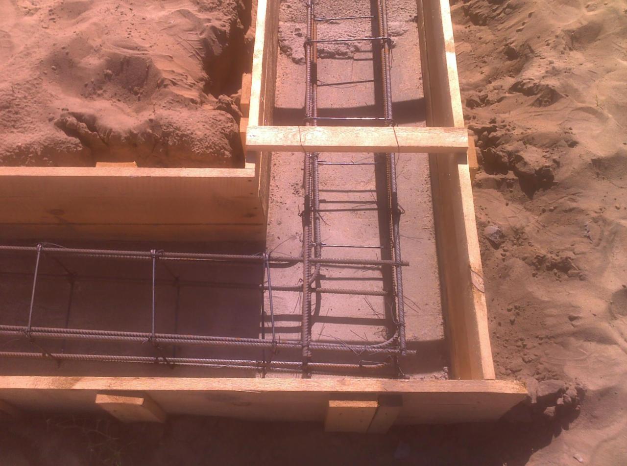 Zbrojenie fundamentów w drewnianym szalunku, widoczne pręty stalowe i piasek w tle.