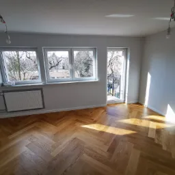 Pusty, jasny pokój z nową podłogą ułożoną w jodełkę, świeżo pomalowanymi ścianami i grzejnikiem. Okna z widokiem na drzewa, a na suficie wiszące przewody elektryczne bez lamp.