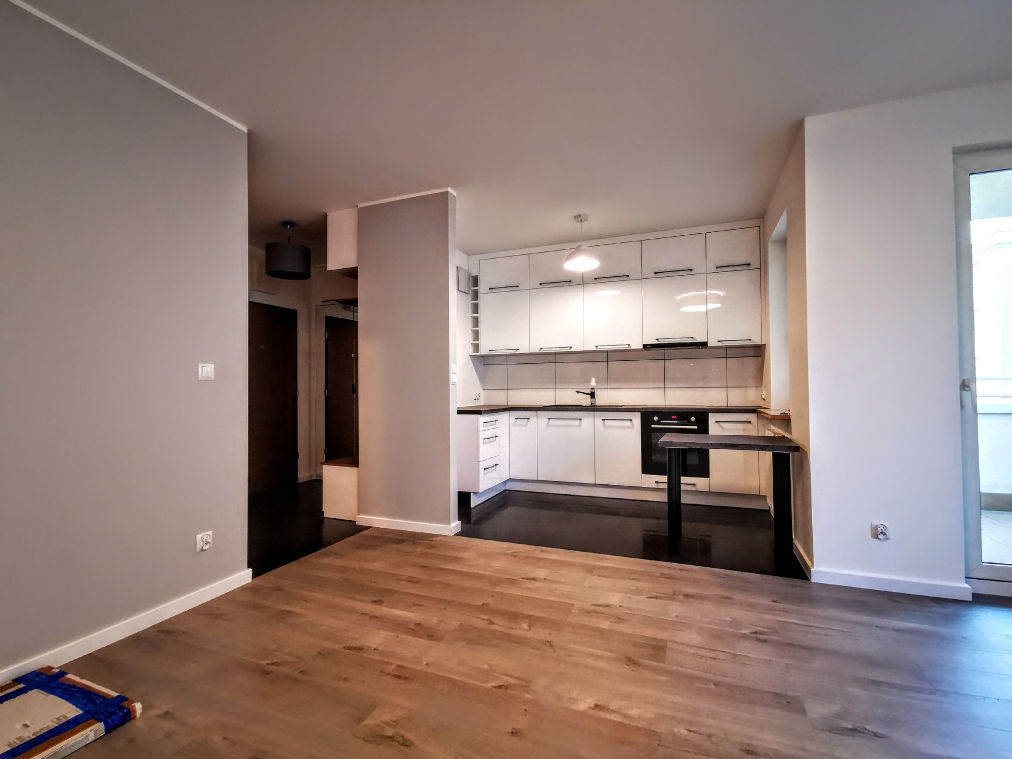 Jasne, przestronne wnętrze mieszkania po remoncie: parkiet w odcieniach brązu, szare ściany, biała zabudowa kuchenna z czarnym blatem i oświetleniem punktowym, widok na przedpokój z ciemną podłogą.