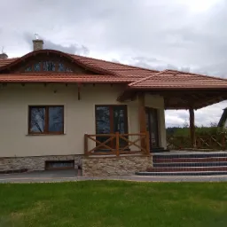 Jasny dom jednorodzinny z czerwoną dachówką, drewnianą werandą i schodami wyłożonymi kostką, widok z trawnika w pochmurny dzień.