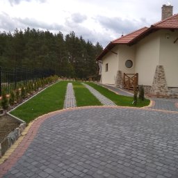 USŁUGI OGÓLNOBUDOWLANE WZNOSZENIE BUDYNKÓW ORAZ REMONTY - Nowoczesny dom z beżową elewacją, czerwoną dachówką i brukowanym podjazdem z dwoma pasami trawy, ogrodzony czarnym metalowym płotem i obsadzony tujami.