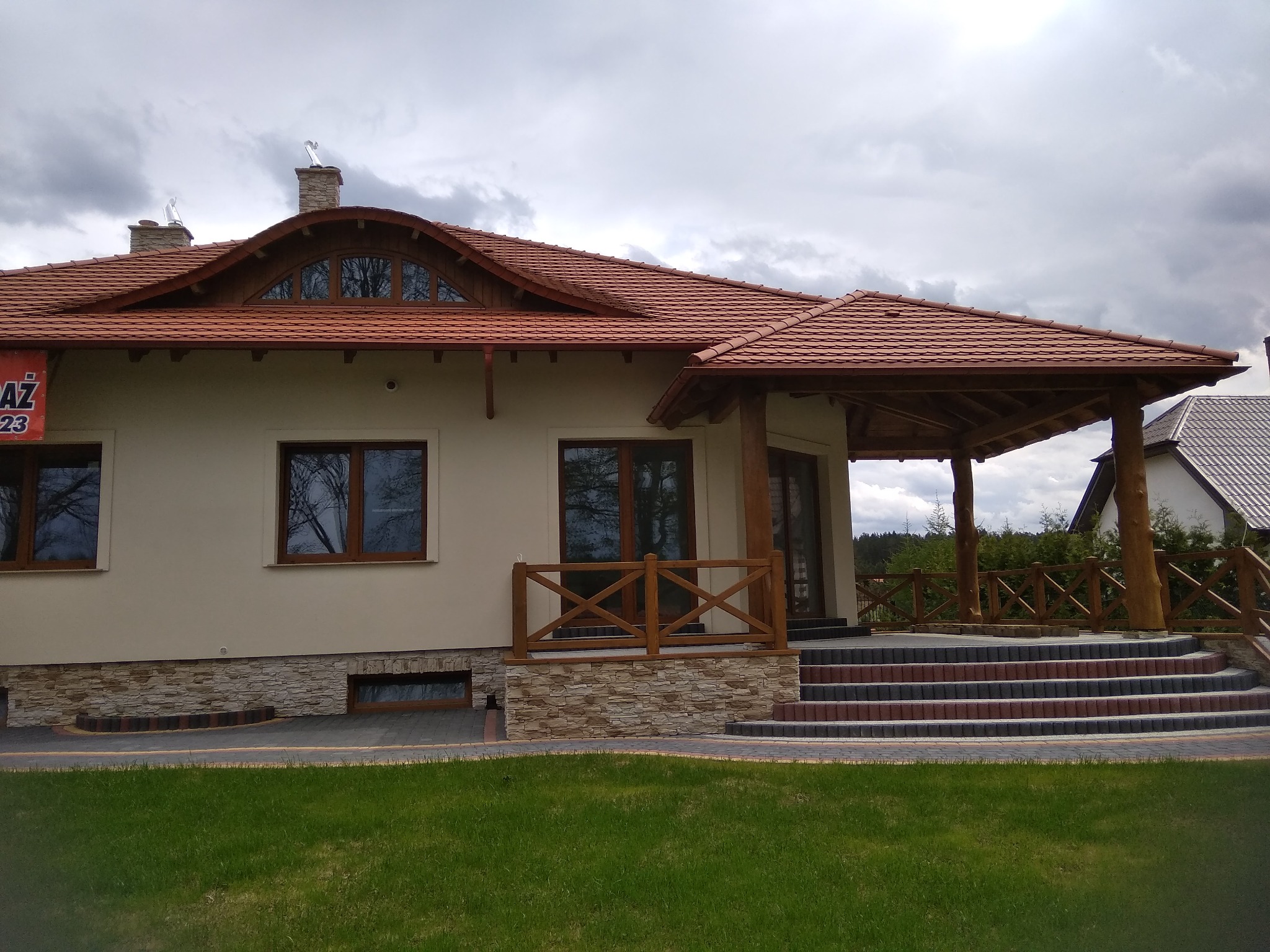 Jasny dom jednorodzinny z czerwoną dachówką, drewnianą werandą i schodami wyłożonymi kostką, widok z trawnika w pochmurny dzień.