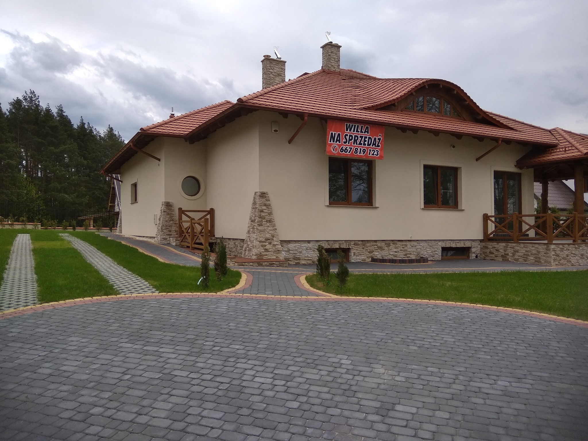 Gotowy, parterowy dom z beżową elewacją, czerwoną dachówką i brukowanym podjazdem, na ścianie baner 'Willa na sprzedaż' z numerem telefonu.