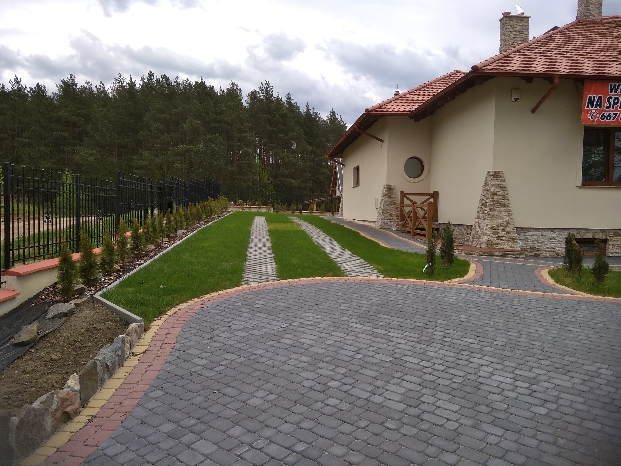 Nowoczesny dom z beżową elewacją, czerwoną dachówką i brukowanym podjazdem z dwoma pasami trawy, ogrodzony czarnym metalowym płotem i obsadzony tujami.