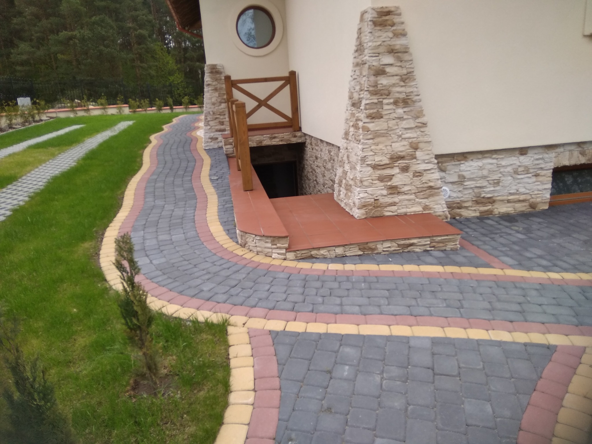 Wejście do piwnicy w domu jednorodzinnym, z drewnianą barierką i schodami wyłożonymi czerwoną terakotą, otoczone ozdobną ścieżką z kostki brukowej w kolorach szarym, czerwonym i żółtym, prowadzącą...