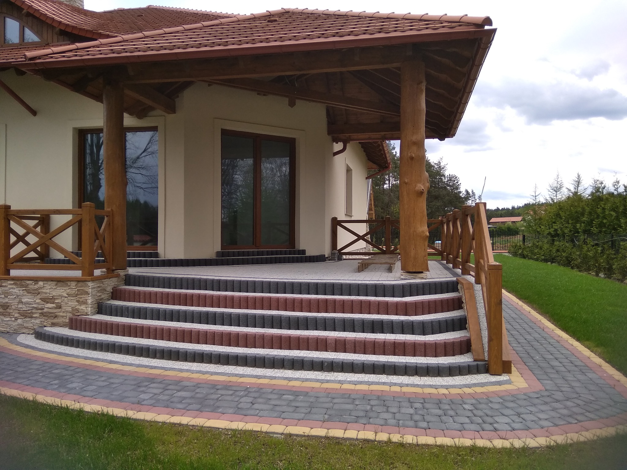 Elewacja domu z drewnianym zadaszeniem wspartym na pniu, schody wejściowe wyłożone kostką w trzech kolorach, chodnik z kostki brukowej prowadzący do domu.