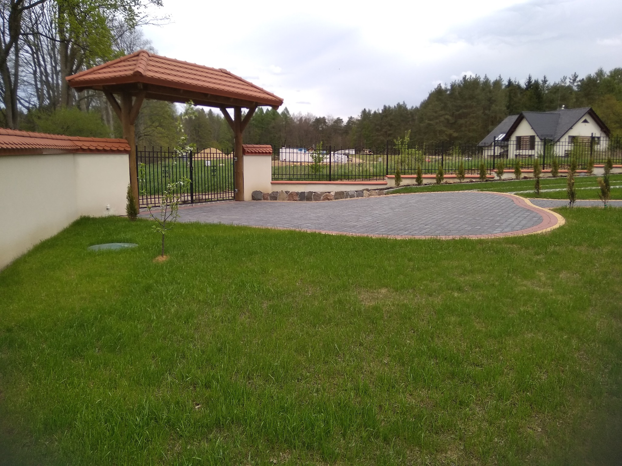 Wjazd na posesję z bramą wjazdową z drewnianym zadaszeniem i metalowym ogrodzeniem na podmurówce z kamienia, wyłożony kostką brukową, z widokiem na dom w tle.