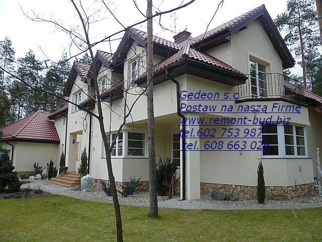 Gotowy, dwukondygnacyjny dom jednorodzinny z balkonem, jasną elewacją i czerwoną dachówką, otoczony zielonym trawnikiem i drzewami.