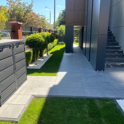 Jacek Domagała - Nowoczesny ogród z równo przyciętymi krzewami, szarą nawierzchnią z płyt, białymi kamykami i elegancką zabudową śmietnikową w kolorze antracytowym.