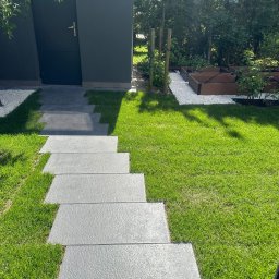 Jacek Domagała - Stopniowa ścieżka z szarych płyt prowadząca przez soczysty trawnik do szarego domku narzędziowego, obok rabaty z białym żwirem i drewnianymi skrzyniami na rośliny.