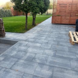 Jacek Domagała - Szara kostka brukowa ułożona na posesji, prowadząca od schodów budynku do trawnika z młodymi drzewkami, widoczna drewniana paleta i skrzynia na kółkach.