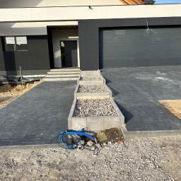 Jacek Domagała - Nowoczesny podjazd z szarej kostki brukowej ułożonej wzorem jodełki, z trzema stopniami z kamieniami ozdobnymi, prowadzący do domu z grafitową bramą garażową i elewacją w kolorze złamanej bieli...