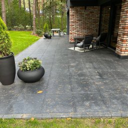 Jacek Domagała - Nowoczesny taras wyłożony szarą kostką z widocznymi igłami sosnowymi, obrzeża z kostki, donice z roślinami ozdobnymi, meble ogrodowe i fragment budynku z cegły w tle.