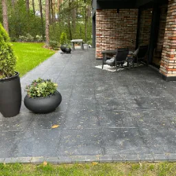 Nowoczesny taras wyłożony szarą kostką z widocznymi igłami sosnowymi, obrzeża z kostki, donice z roślinami ozdobnymi, meble ogrodowe i fragment budynku z cegły w tle.