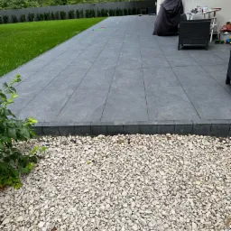 Wyłożony szarymi płytami taras w Raszynie, z obrzeżem z kostki i jasnym żwirem. W tle trawnik i wysokie drzewa.