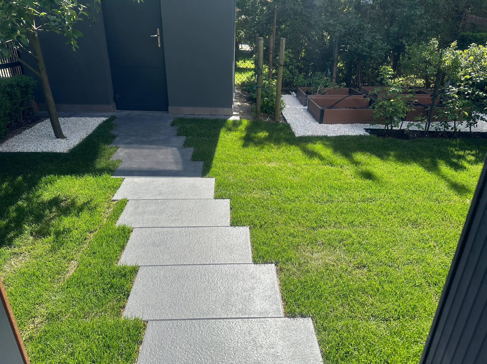 Stopniowa ścieżka z szarych płyt prowadząca przez soczysty trawnik do szarego domku narzędziowego, obok rabaty z białym żwirem i drewnianymi skrzyniami na rośliny.
