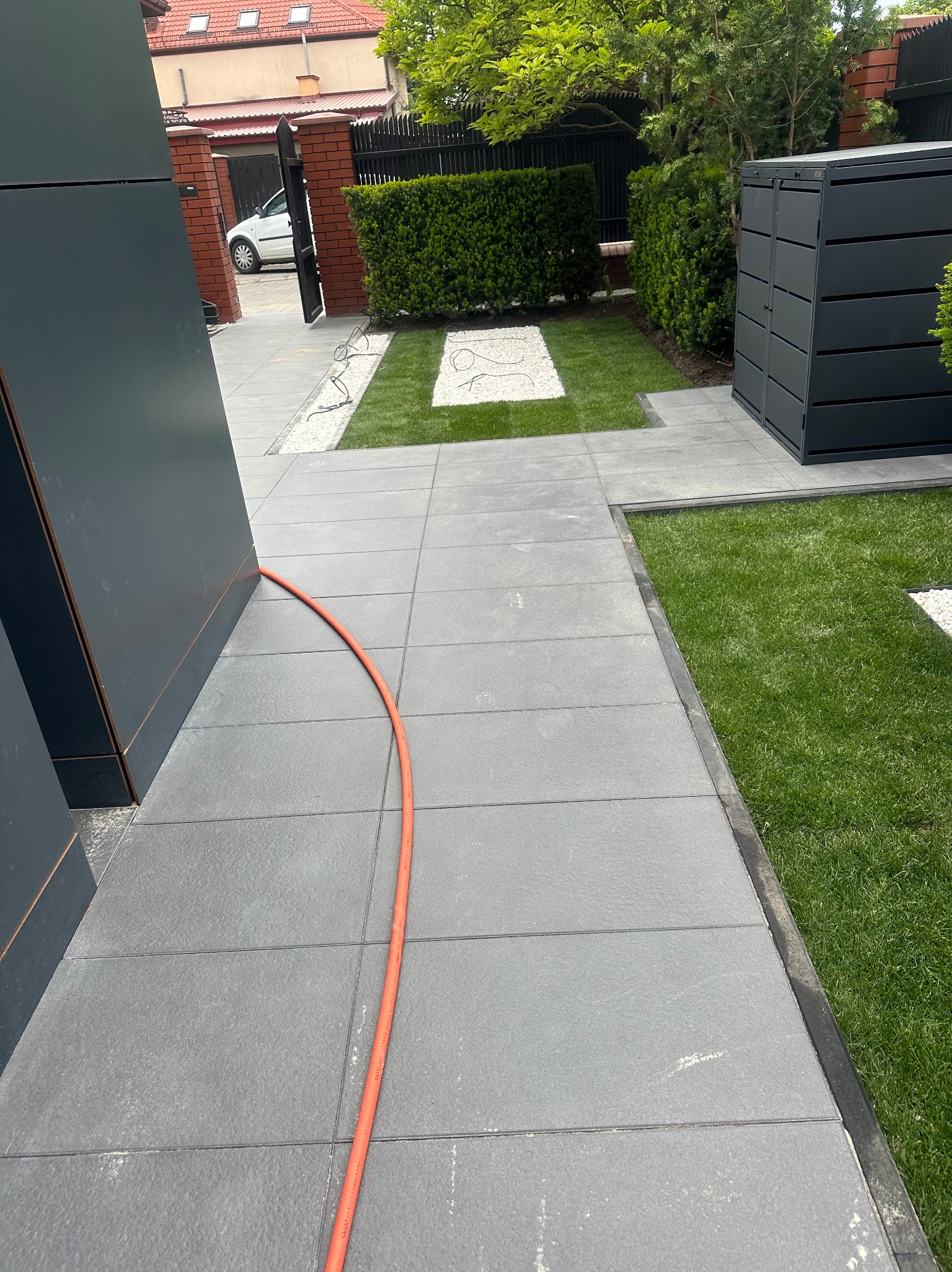 Nowoczesna aranżacja przestrzeni przydomowej z szarych, kwadratowych płyt betonowych, zielonym trawnikiem i białymi kamykami, obok czarnej zabudowy gospodarczej i żywopłotu.