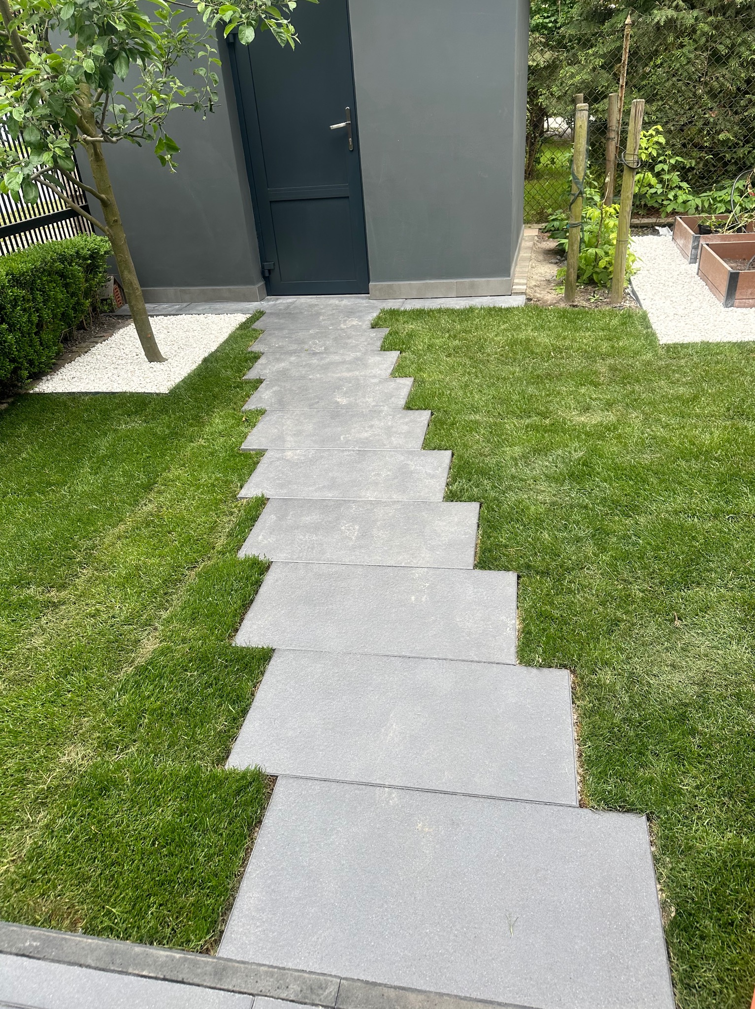Ścieżka z płyt betonowych prowadząca do ciemnoszarego budynku w ogrodzie, obok trawnik, drzewo i białe kamyki.