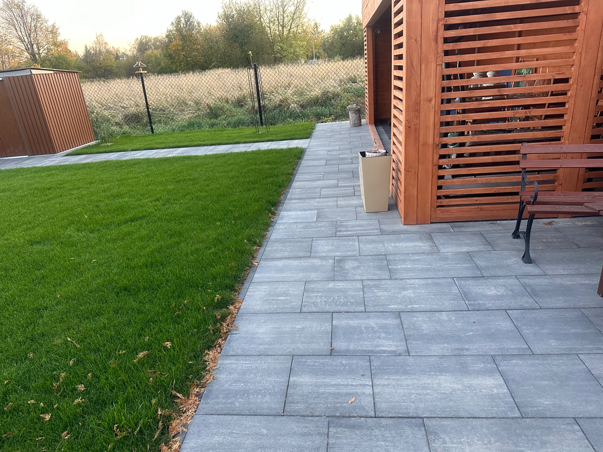 Ułożona szara kostka betonowa prowadzi wzdłuż trawnika do drewnianej altany z ławką, widoczny również brązowy domek narzędziowy w tle.