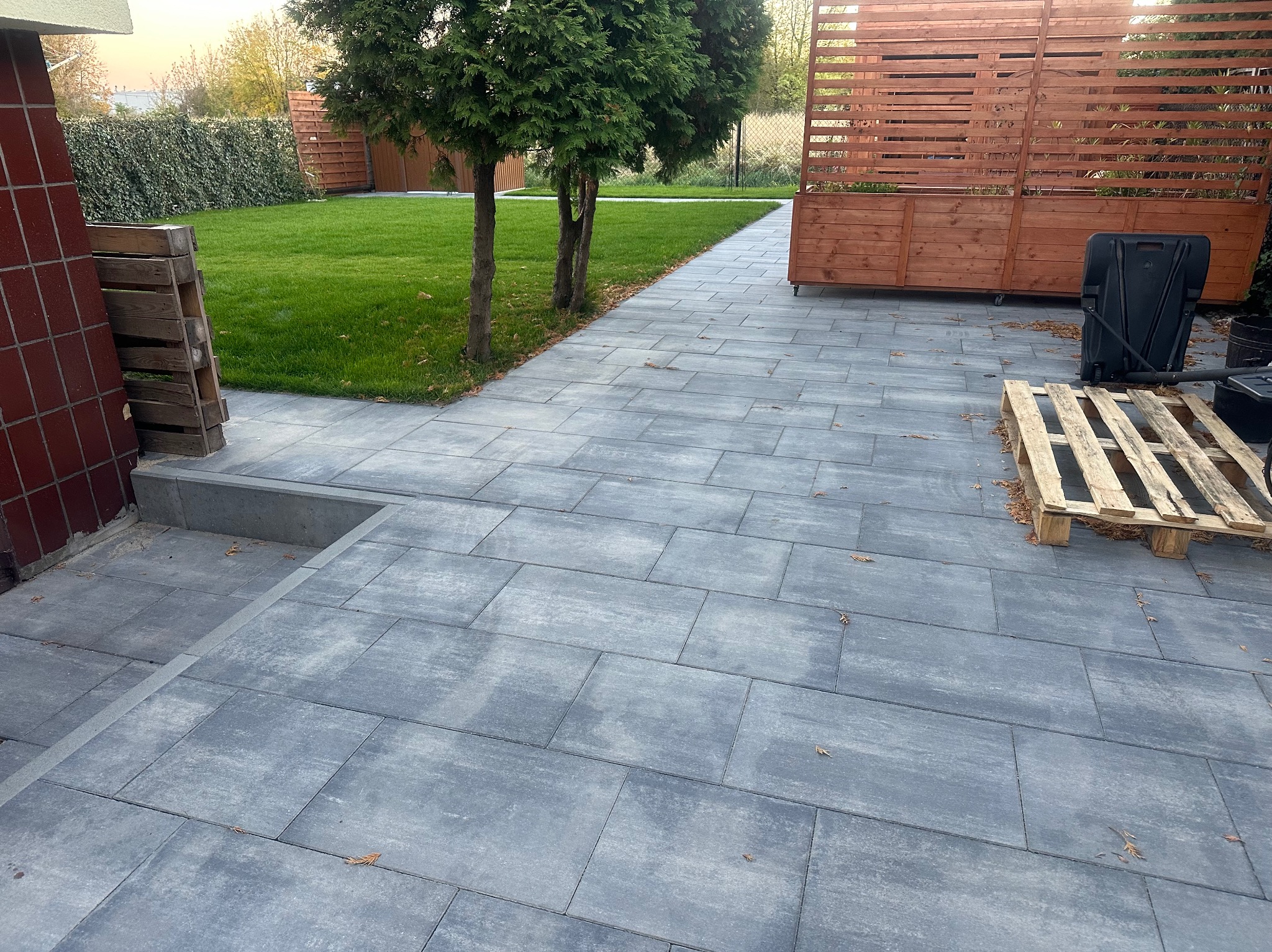 Szara kostka brukowa ułożona na posesji, prowadząca od schodów budynku do trawnika z młodymi drzewkami, widoczna drewniana paleta i skrzynia na kółkach.