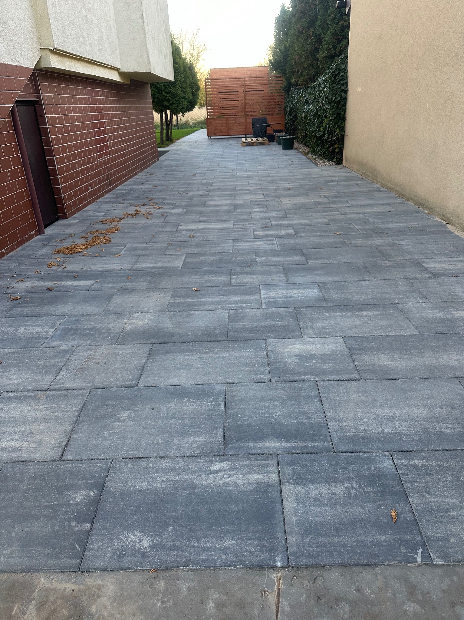 Szara, prostokątna kostka betonowa ułożona na podjeździe w Raszynie, widok z perspektywy osoby stojącej na końcu podjazdu, w tle drewniana konstrukcja ogrodowa i zielone tuje.