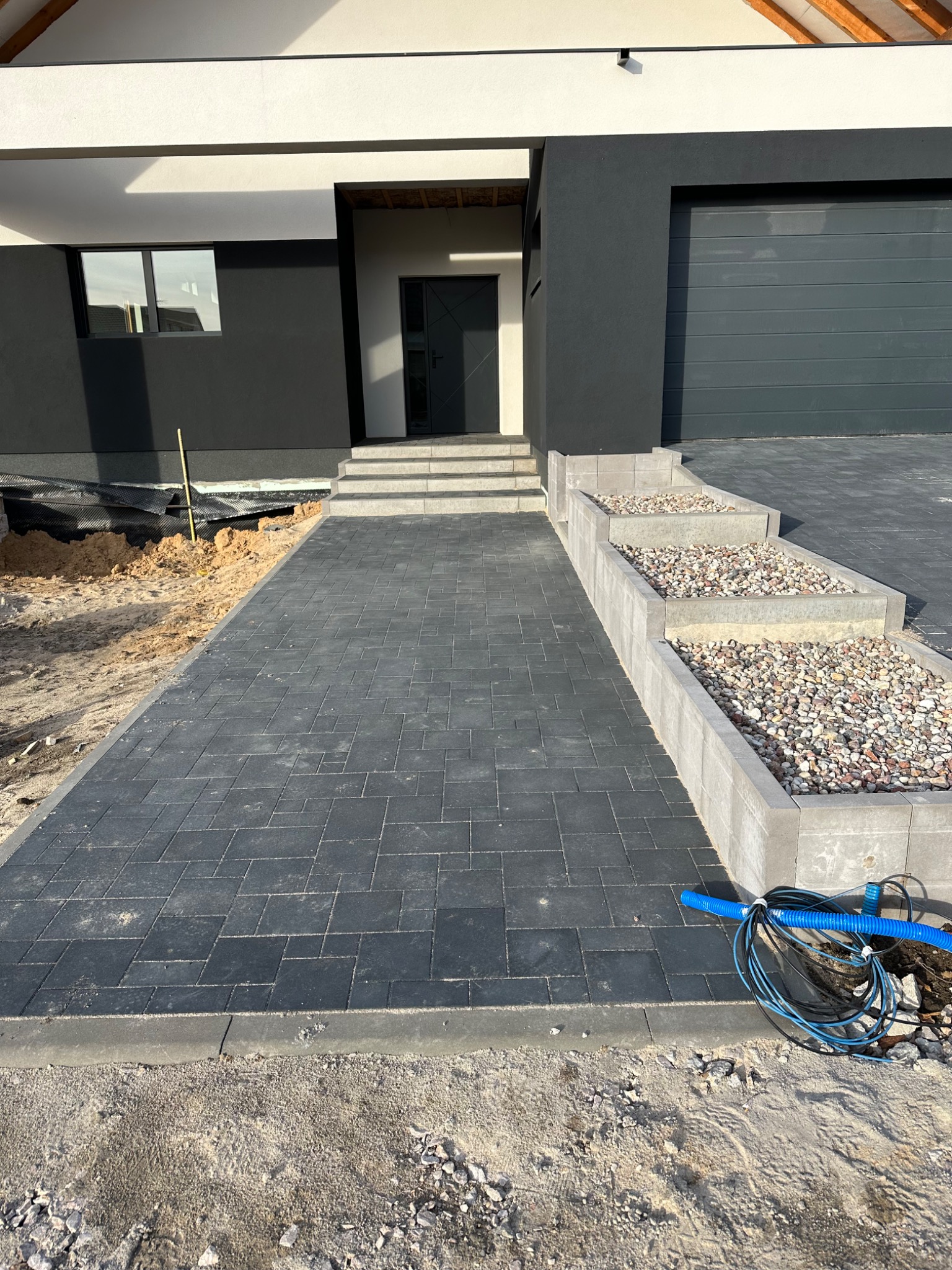 Nowoczesny podjazd z ciemnej kostki betonowej prowadzący do domu z szarą elewacją i garażem, z elementami dekoracyjnymi w postaci podwyższonych rabat wypełnionych drobnym kamieniem, obok widoczne...