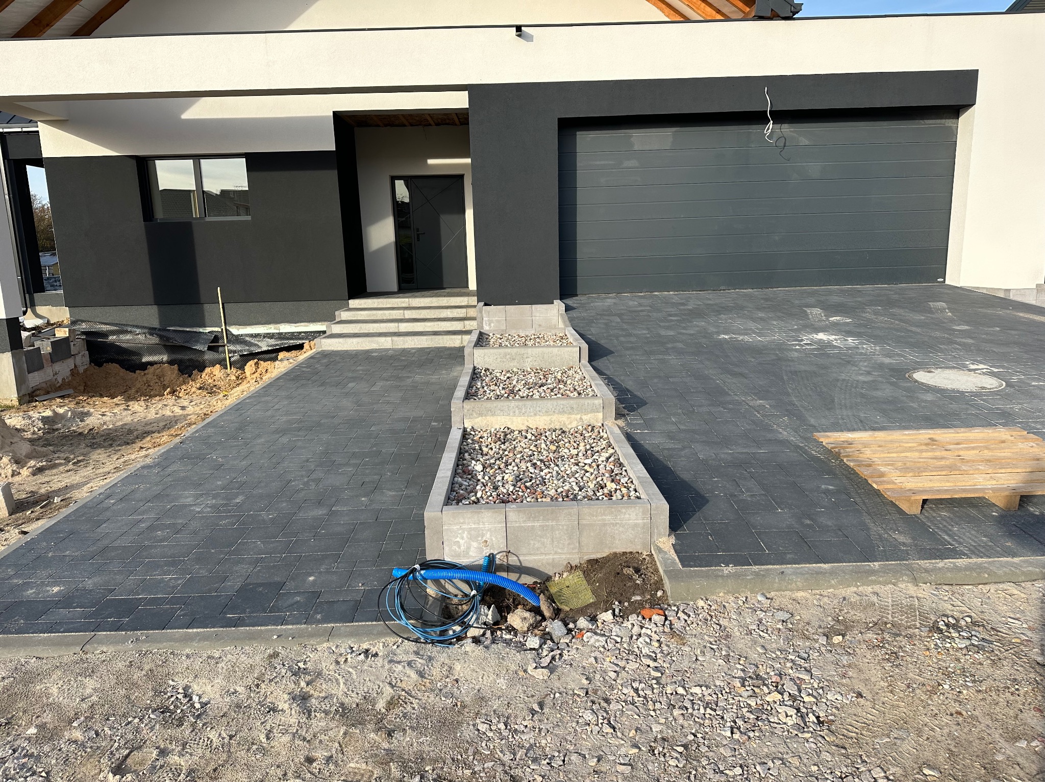 Nowoczesny podjazd z szarej kostki brukowej ułożonej wzorem jodełki, z trzema stopniami z kamieniami ozdobnymi, prowadzący do domu z grafitową bramą garażową i elewacją w kolorze złamanej bieli...