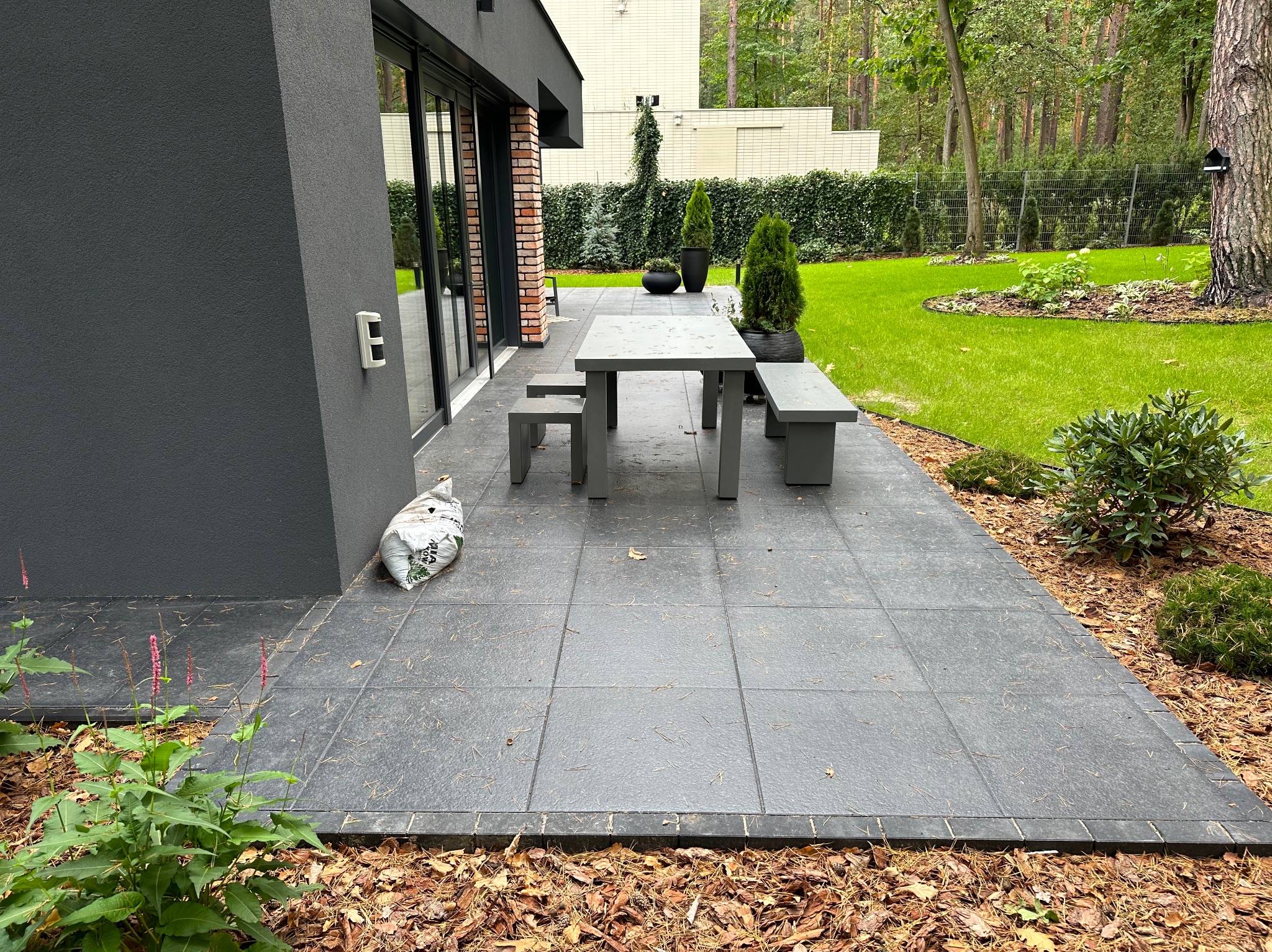 Nowoczesny taras z szarych płyt betonowych, otoczony korą i zielenią, z minimalistycznym zestawem mebli ogrodowych i widokiem na zadbany ogród z tujami i trawnikiem.