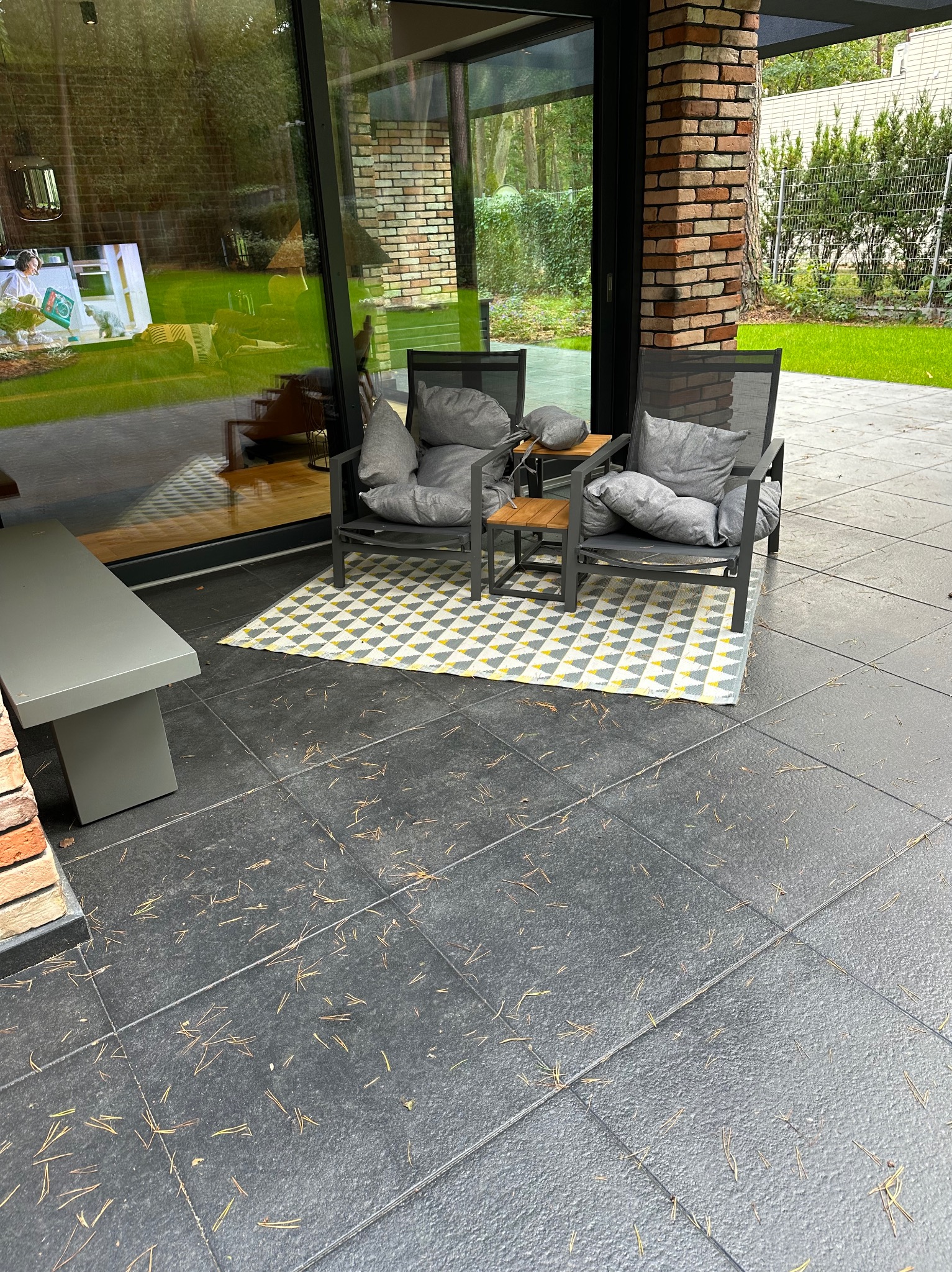 Zewnętrzny taras z ciemnych płyt, wyposażony w dwa metalowe krzesła z szarymi poduszkami, mały drewniany stolik i geometryczny dywanik w odcieniach szarości i żółci; igliwie rozsypane na płytach.