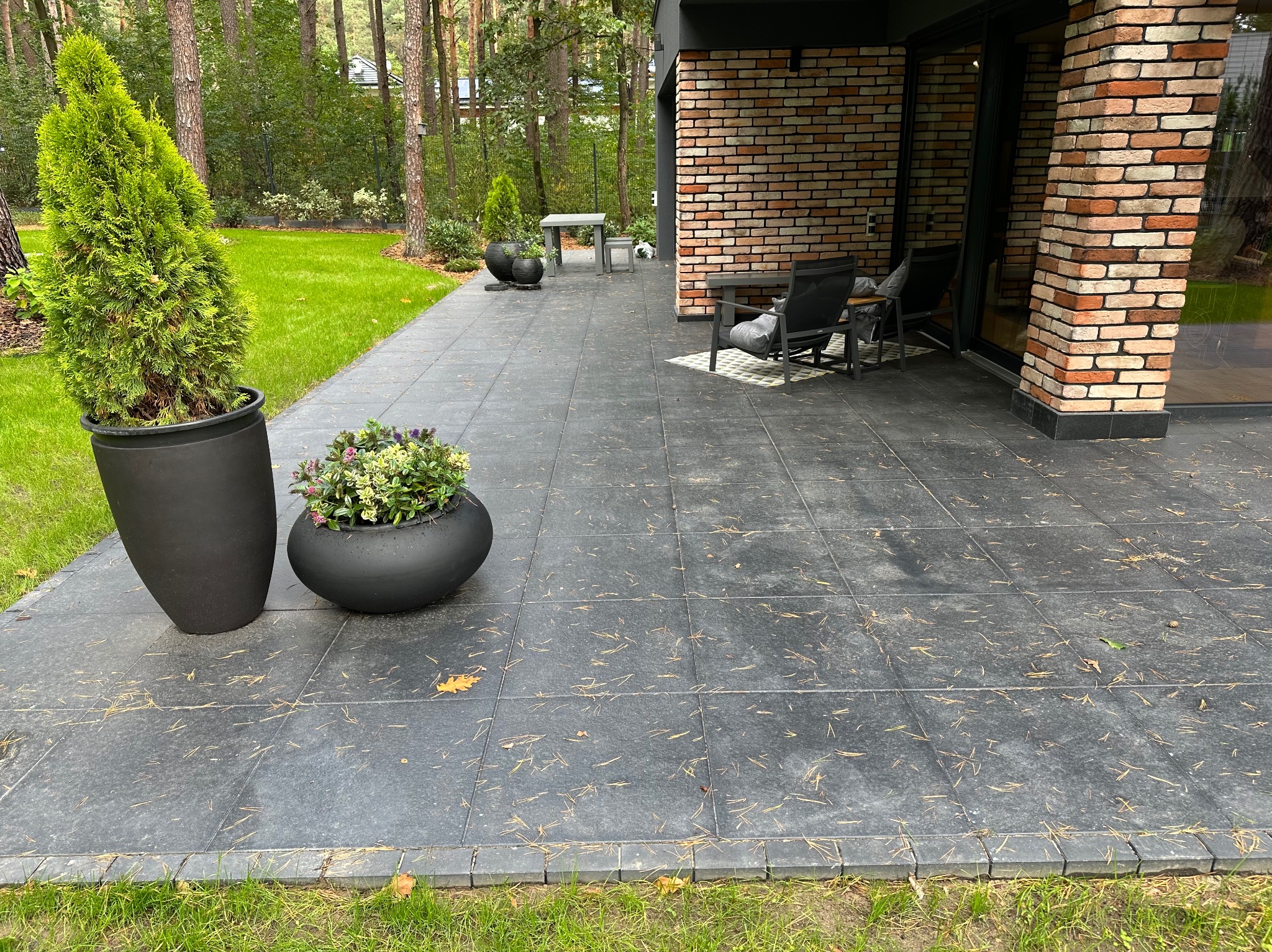 Nowoczesny taras wyłożony szarą kostką z widocznymi igłami sosnowymi, obrzeża z kostki, donice z roślinami ozdobnymi, meble ogrodowe i fragment budynku z cegły w tle.