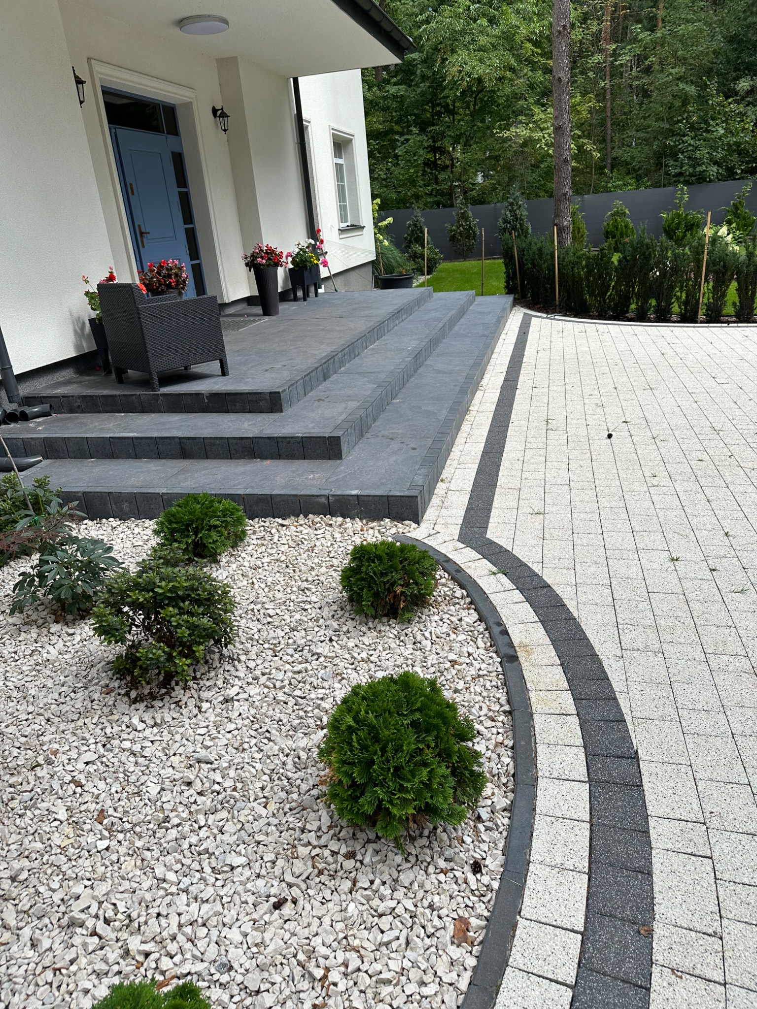Nowoczesny podjazd z jasnej kostki z ciemnymi obrzeżami, kontrastujący z szarymi schodami i białym domem z niebieskimi drzwiami, uzupełniony rabatą z białego kamienia i zielonymi krzewami.