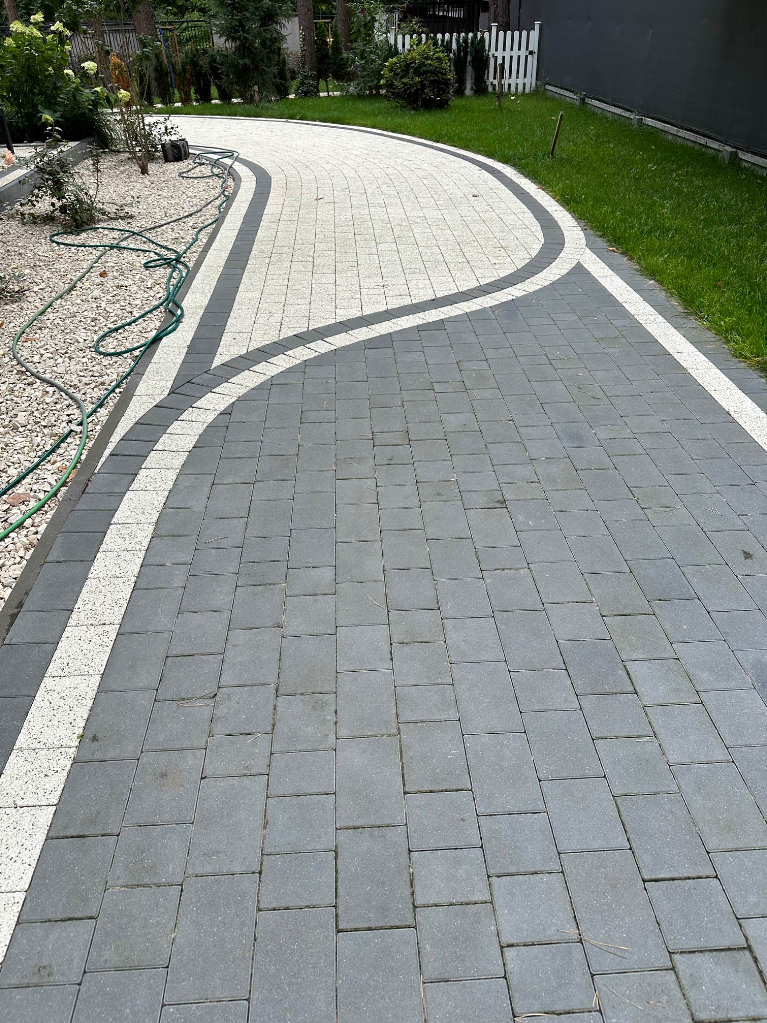 Zakrzywiony chodnik z szarych kostek betonowych z białymi i ciemnoszarymi obrzeżami, otoczony zielonym trawnikiem i kruszywem.