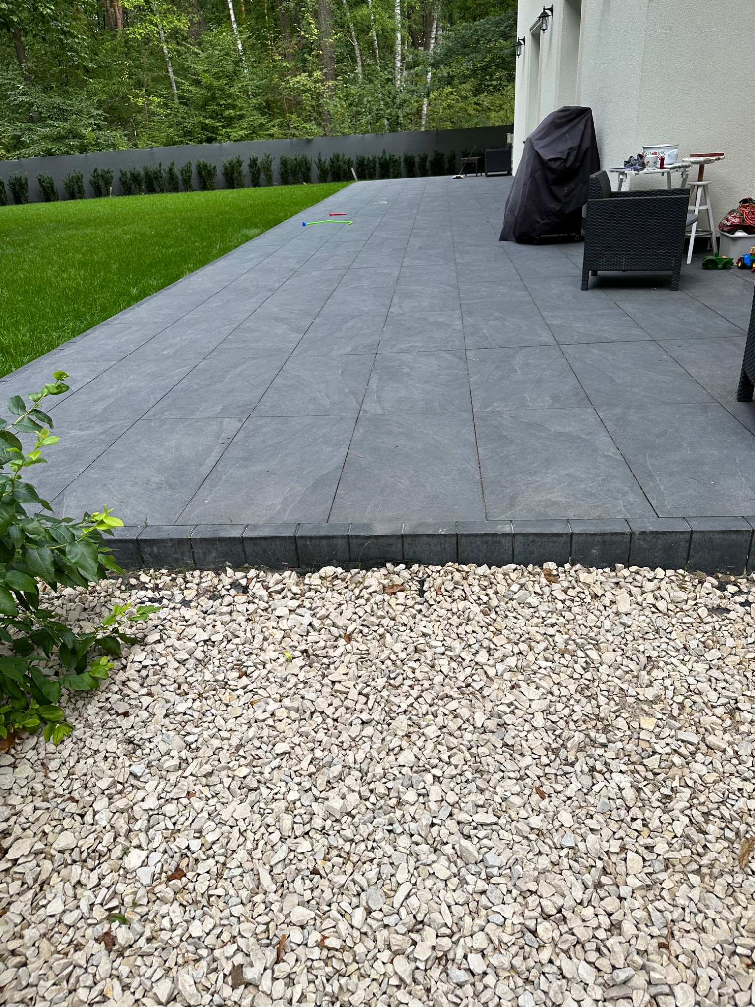 Wyłożony szarymi płytami taras w Raszynie, z obrzeżem z kostki i jasnym żwirem. W tle trawnik i wysokie drzewa.