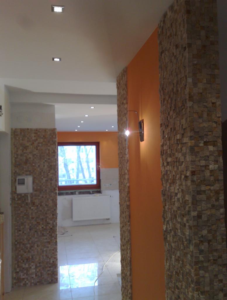 Aranżacja wnętrza: fragment ściany z dekoracyjnym kamieniem w odcieniach beżu i brązu, oświetlona kinkietem, widok na pomieszczenie z oknem i białym grzejnikiem.