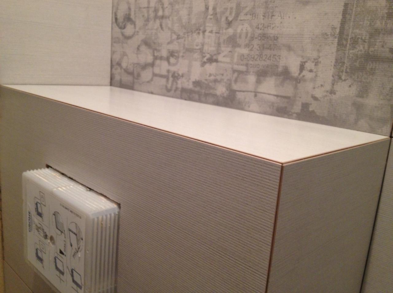 Obudowa WC z białych płytek imitujących drewno, z widocznym przyciskiem spłukującym, na tle ściany z szarym wzorem przypominającym graffiti.